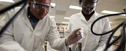 Two students in lab coats and safety goggles performing tests in a lab